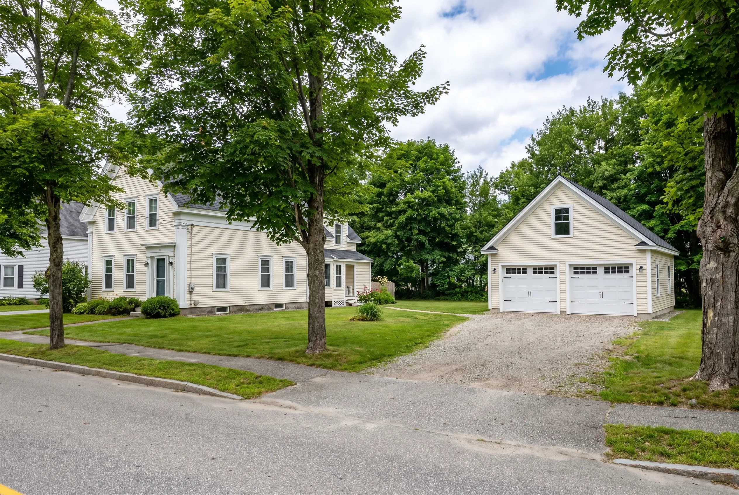 Two-bay detached garage in Gardiner, Maine: White vinyl-sided garage with two white garage doors sits on gravel driveway beside white colonial house, surrounded by mature trees and green lawn.