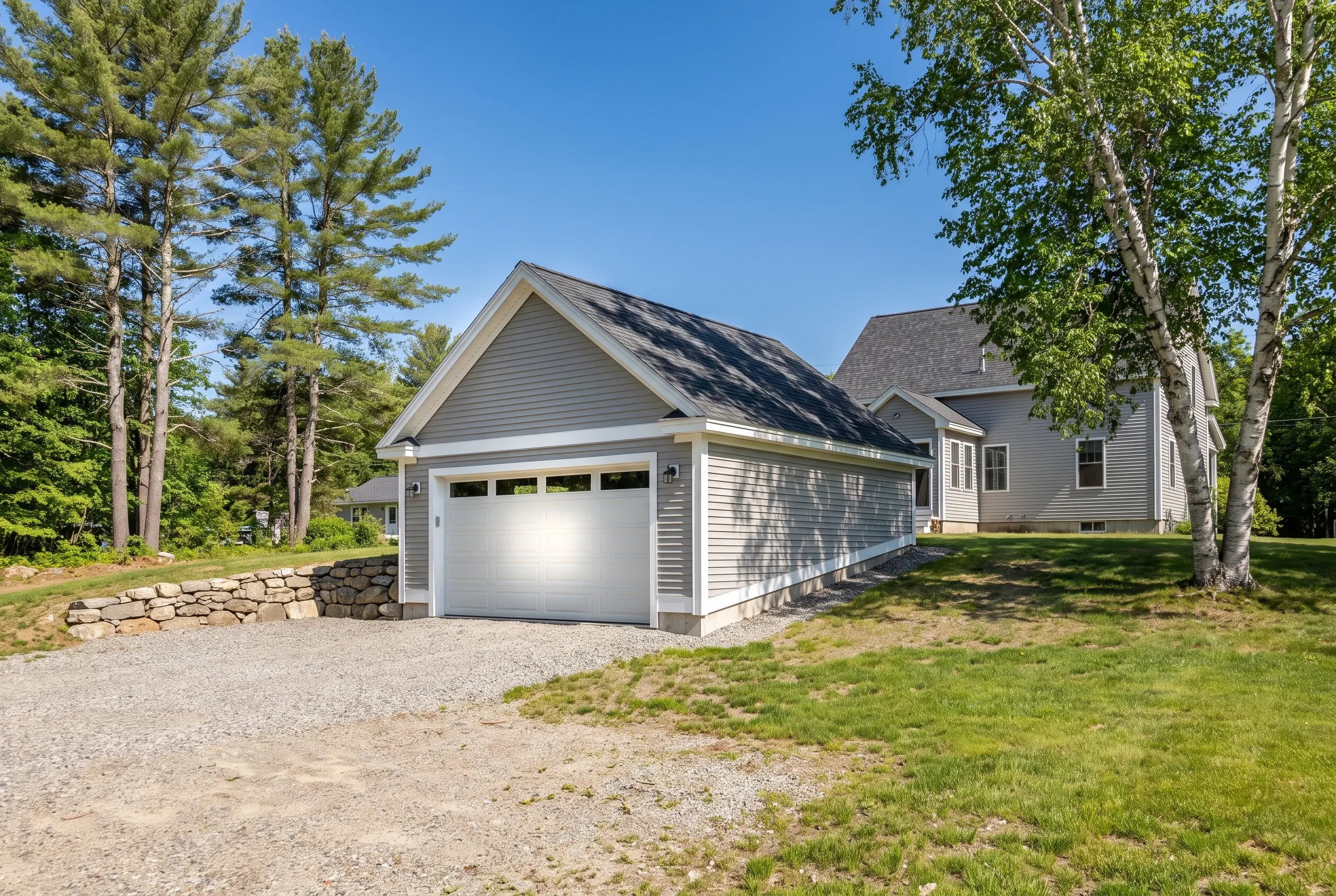 Detached two-bay garage in Hallowell, Maine: White garage with dark roof beside residential home, surrounded by tall trees and stone wall, gravel driveway.