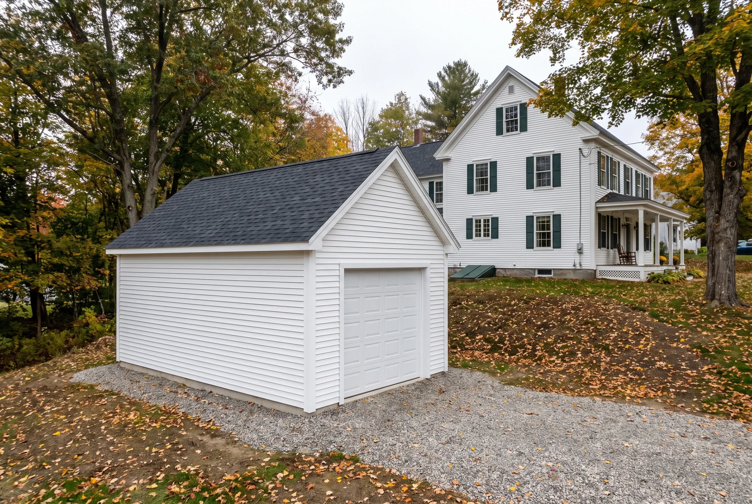Detached two-bay garage in Hallowell, Maine: White single-story garage with gray roof and white garage door on gravel driveway, adjacent to white two-story colonial house surrounded by fall foliage.