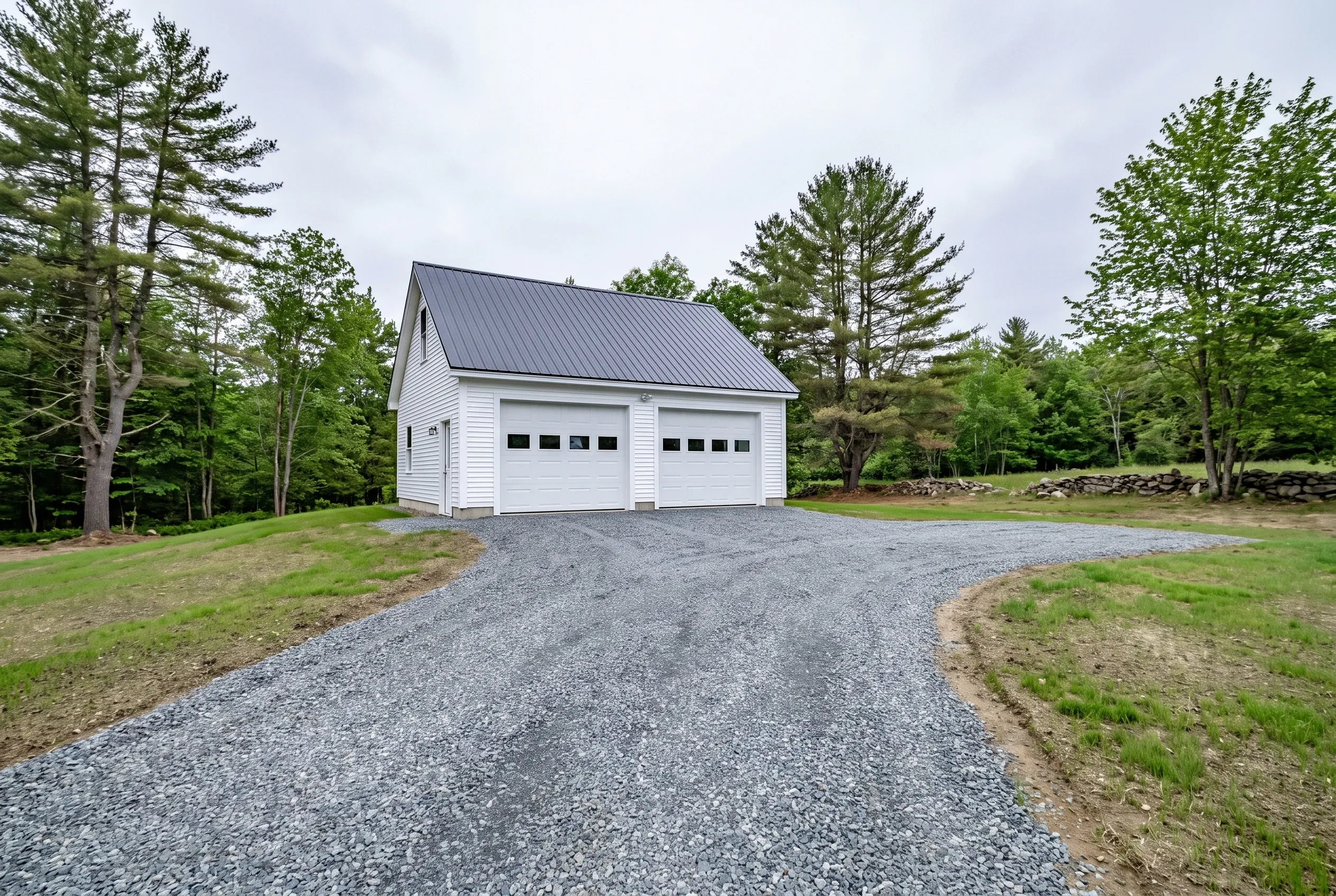 Detached two-bay garage in Vassalboro, ME: White vinyl sided garage with metal roof on gravel driveway surrounded by trees and grass.